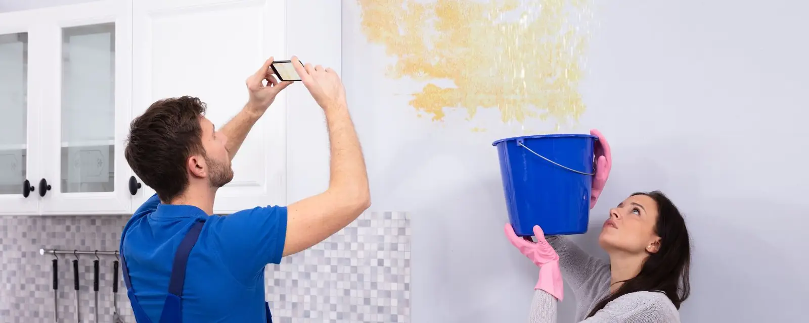 Man photographing water stained wall damage while a woman holds a bucket beneath the leak